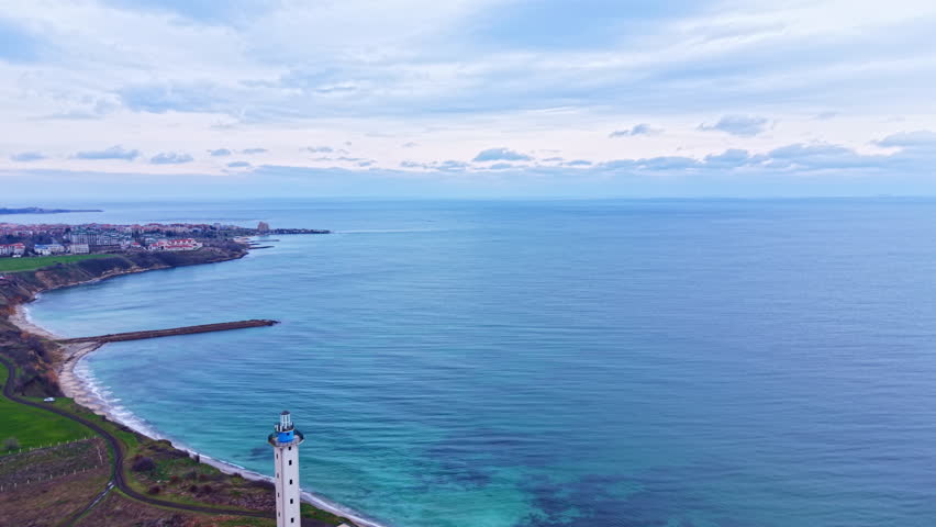 A lighthouse stands near the shore with a view of the ocean and a small town in the distance. Clouds cover the sky above the calm water.