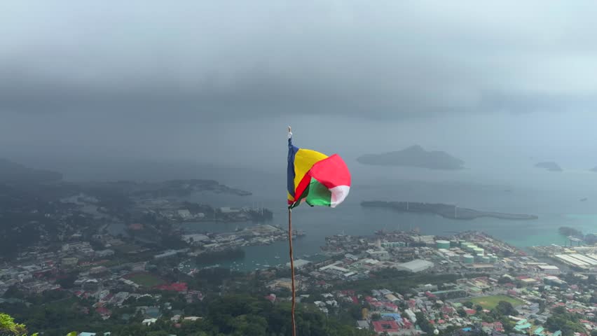 Seychelles flag floating above hill, beneath town Victoria and port area, Mahe, Seychelles 25fps 002
