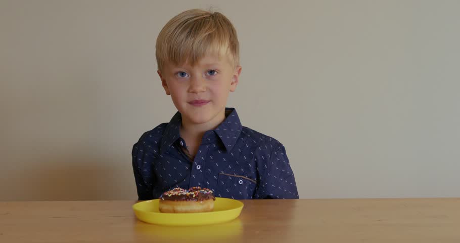 A slow-motion shot of a young boy holding a chocolate-glazed donut with sprinkles in front of him, happily smelling it and smiling.