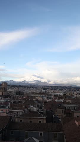 Vertical screen snowy Mount Etna rising above the city of Catania with calm blue sky and soft clouds creating a peaceful southern atmosphere blending urban texture with natural volcanic landscape