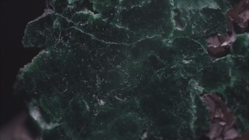 Microscopic macro image of a polished malachite specimen from the Kamenushinskoe copper deposit, captured using an optical microscope with resolution up to 2.68 micrometers and field of view 15 mm. The surface displays concentric banding with zones of emerald green, bluish-green, and pale green, reflecting variations in copper carbonate composition during sequential crystallization. Microfractures intersect the polished plane, some partially filled with fine-grained secondary minerals. Dark opaque inclusions—identified as chalcopyrite, hematite, and minor pyrite—are distributed irregularly and contrast strongly with the translucent host. Surface texture is smooth but reveals topographic variation due to differential hardness and polishing wear; individual crystal domains are resolvable at this resolution. Lighting is even and directional, producing soft specular highlights that emphasize relief along fracture edges and inclusion boundaries without artificial enhancement. No digital filters or compositing were applied. This documentation serves as a source of scientific knowledge for the study of secondary mineral textures, microfracture-filling processes, and paragenetic relationships in supergene copper deposits. It supports educational materials in economic geology and mineral identification for visual analysis of diagnostic features such as banding symmetry, inclusion morphology, and reflectance behavior. Used in university-level courses for the study of ore genesis, weathering profiles, and microscopic textural evolution. Such imagery aids in understanding the dynamics of the living world of Earth through surficial geochemical transformations that generate economically and aesthetically significant minerals. Through precise observation and imaging, this work reinforces empirical methods in earth science research and encourages deeper engagement with the structural complexity of natural materials 