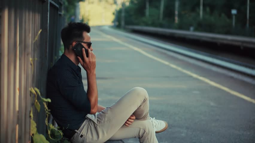 Businessman Talking On Smartphone While Sitting On Railway Platform, Passenger Waiting For Train. Mobile Tech In Travel, Urban Lifestyle And Remote Work, Professional Manager Using Cellphone. Railway