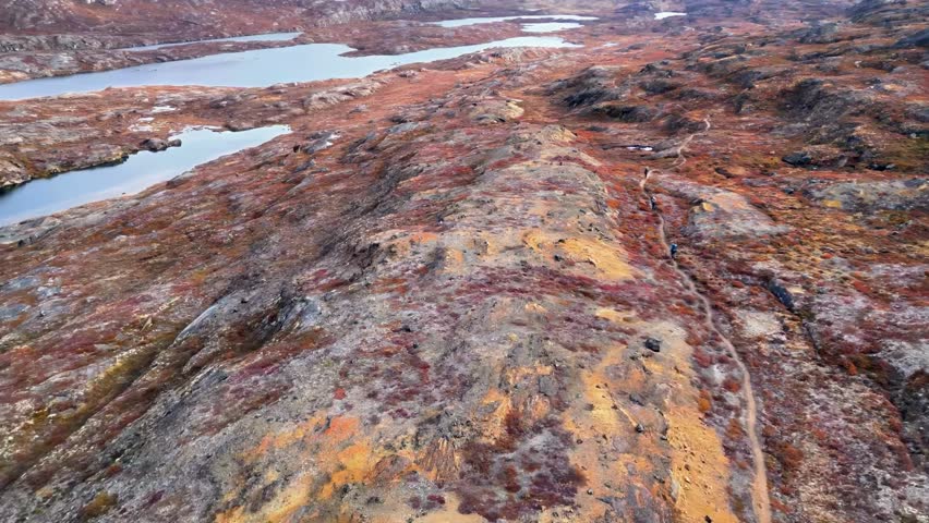 Aerial view of the rugged Greenlandic terrain showcasing vibrant autumn tundra colors, rocky outcrops, and serene glacial lakes with a narrow path for hikers