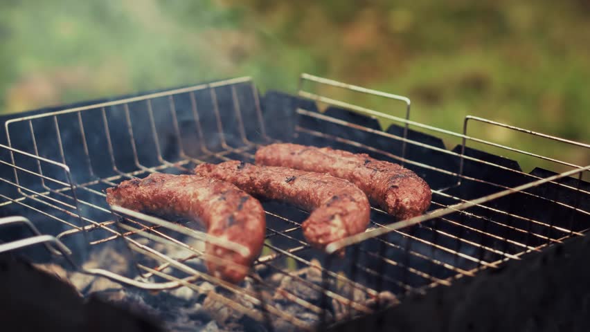 Sausages Grilling On Metal Wire Rack Over Hot Coals In Mangal, Meat Bratwurst Roasting On Open Fire. Barbecue Preparation. Outdoor Cooking And Grilling. Summer Garden Party. Barbeque Meal Picnic Food