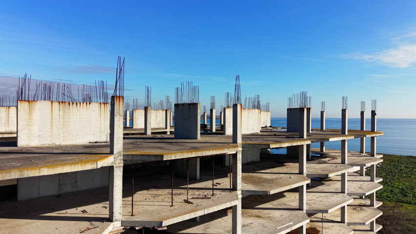 High angle shot showing the top floors of an abandoned or unfinished building with many concrete pillars and exposed rebar near the sea. Aerial view.