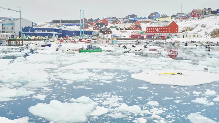 A scenic view of a coastal town in Greenland, featuring boats navigating through icy waters with vibrant buildings on the shore