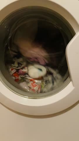 rotating laundry visible through washing machine porthole, mixed garments tumbling with water and suds, motion and blur emphasize active cleaning cycle Video taken on mobile device.