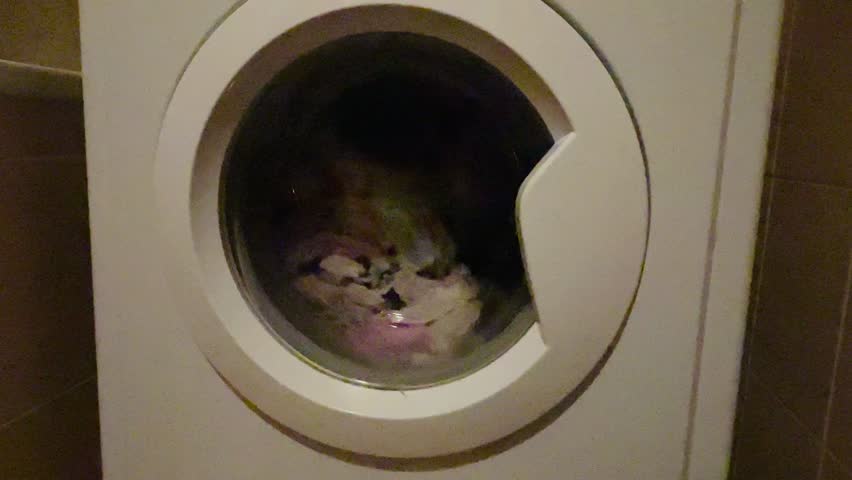 front drum showing spinning clothes inside, vivid pink and white textiles blur through circular glass, suds and water movement visible, energetic tumble Video taken on mobile device.