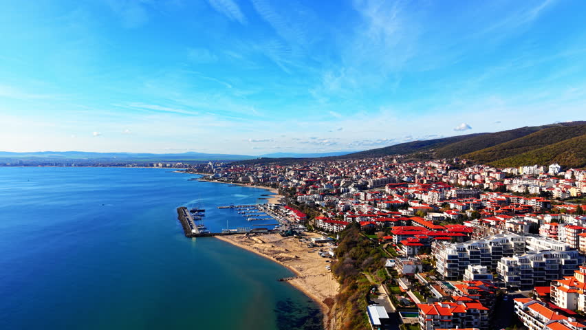 Seaside town with red-roofed buildings stretching along the coastline under a blue sky. Panoramic aerial view of coastal city and harbor.