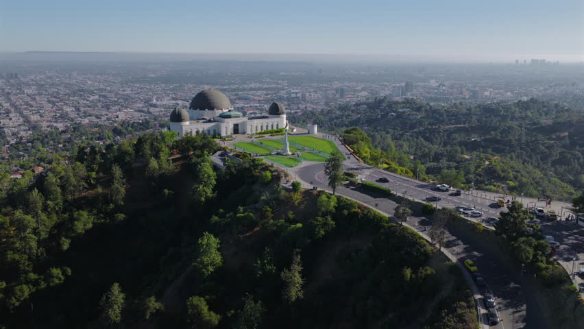 Aerial view of Griffith Observatory above Los Angeles California with city skyline, green hills, roads and traffic. Iconic landmark captured by drone in clear daylight conditions.