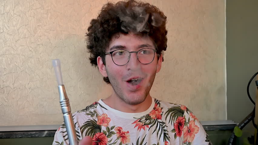 Izmir,Turkey,august 10, 2025. A young man with curly hair and glasses smoking a hookah. He holds the pipe, inhales the flavored vapor, and then exhales a cloud of smoke. Slow motion.