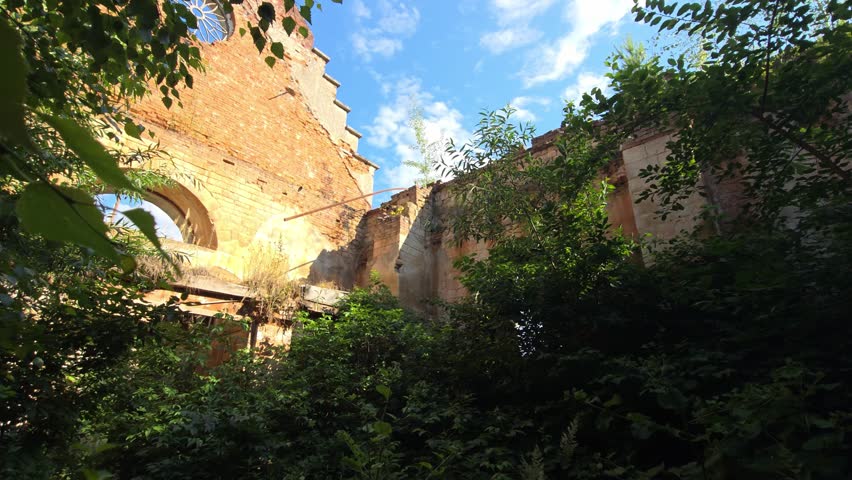 Abandoned red brick church ruins with an arched entrance and circular window sit overgrown with green foliage and trees under a clear blue sky, capturing a sense of lost history