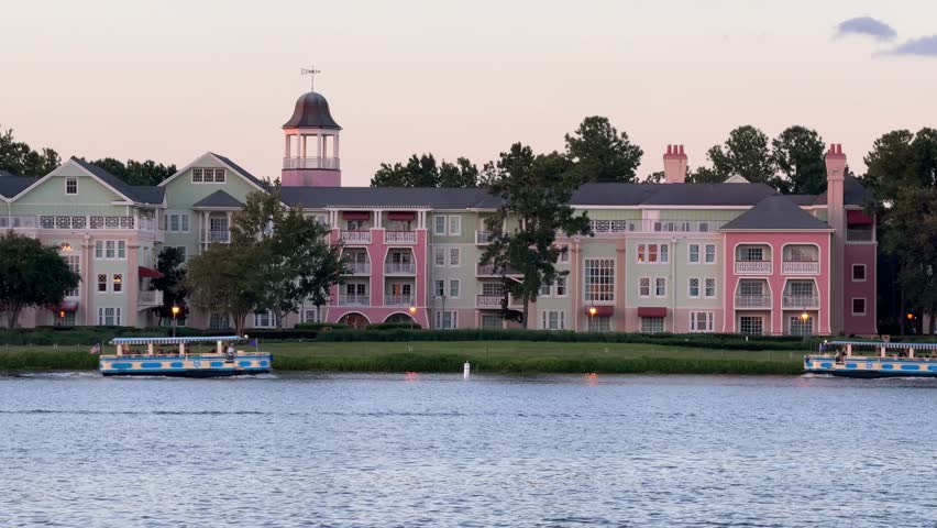 Pastel resort buildings stand along a calm lake while two tour boats cruise in front. Soft evening light creates a peaceful waterfront vacation scene in Orlando, Florida.