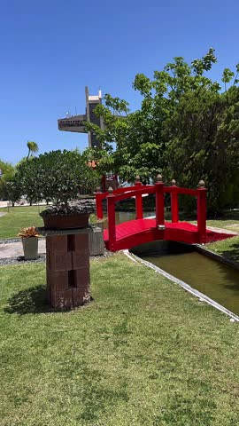 A bright red wooden bridge crosses a calm pond within a carefully landscaped garden, surrounded by lush greenery, bonsai-style plants, and open blue skies, creating a serene and balanced outdoor environment.