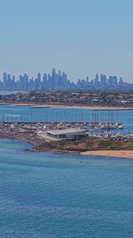 Drone aerial view of Sandringham Beach and Port Phillip Bay in Melbourne, Australia, showing sandy shoreline, beachgoers, calm blue water, marina, coastal vegetation, and the city skyline on a clear sunny day.