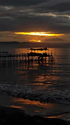 Silhouetted wooden pier on a tropical beach at sunset with dramatic cloudy sky and golden reflections on the sea waves.
