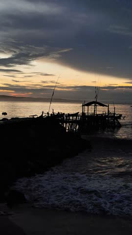 Silhouetted wooden pier on a tropical beach at sunset with dramatic cloudy sky and golden reflections on the sea waves.