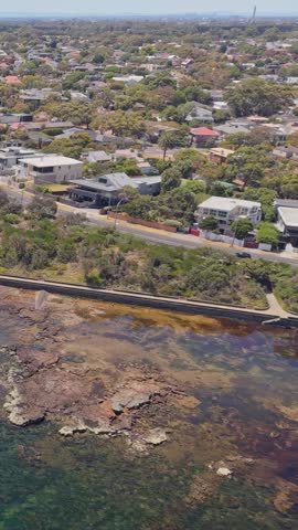 Drone flyover along the Black Rock coastline in Melbourne, Australia, featuring the Bay Trail coastal path, rocky shoreline, clear blue water, suburban homes, and distant city skyline on a sunny day.