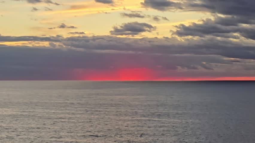 Vivid red sunset glowing on the horizon over calm ocean waters with dramatic clouds, creating a cinematic and atmospheric seascape at dusk.