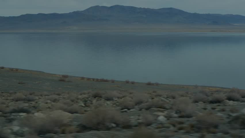 Vast Pyramid Lake in Nevada at dusk with calm waters, desert shoreline with sparse shrubs under hazy sky, distant blue mountains reflected, concept of serene wilderness and natural tranquility