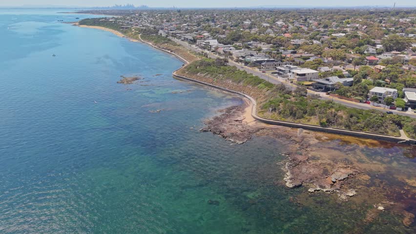 Drone flyover along the Black Rock coastline in Melbourne, Australia, featuring the Bay Trail coastal path, rocky shoreline, clear blue water, suburban homes, and distant city skyline on a sunny day.