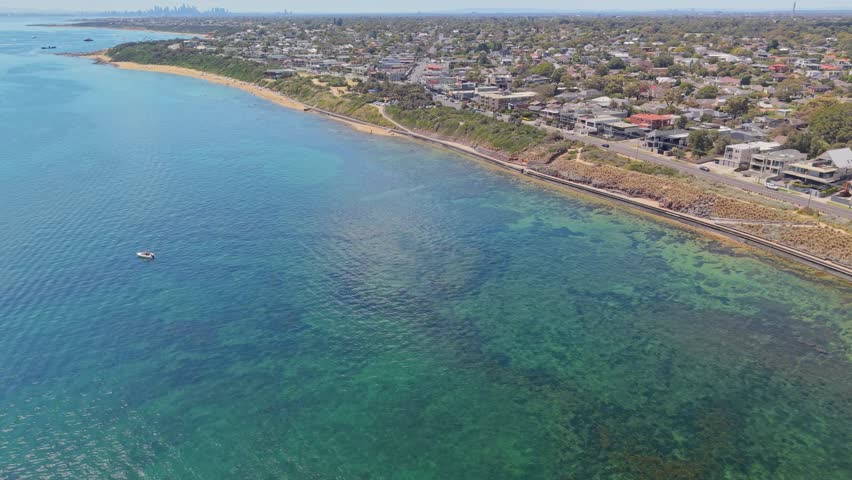 Drone flyover along the Black Rock coastline in Melbourne, Australia, showing the Bay Trail, sandy beaches, clear turquoise water, suburban housing, boats, and distant CBD skyline on a bright summer day.
