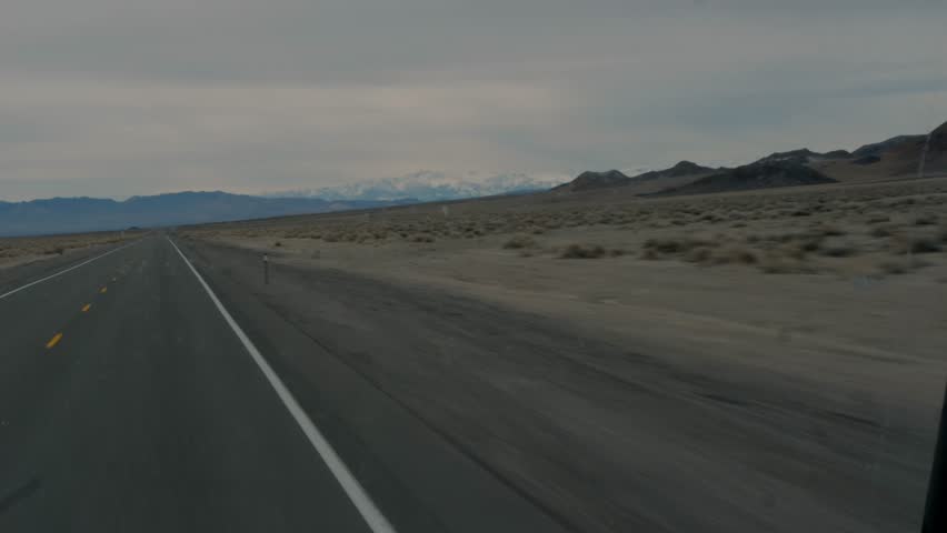 Nevada desert highway under cloudy sky leading to snowcapped mountains, straight empty two-lane road through barren sand and scrub with rocky hills, concept of remote winter road trip solitude
