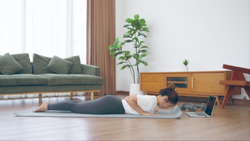 Young Asian woman stretching yoga workout on exercise mat while online training class with computer laptop in living room
