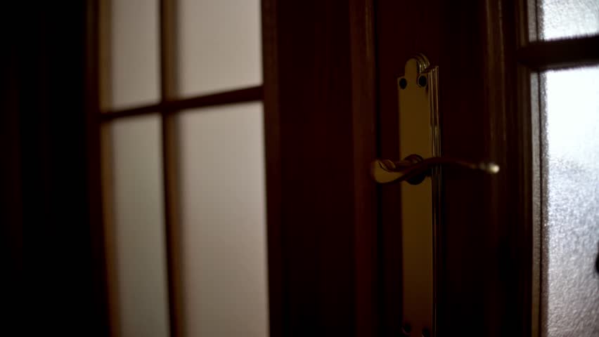 A close-up of a hand turning a brass door handle and pulling open a wooden door into a softly lit living space. The scene conveys home, entry, privacy, and everyday life.