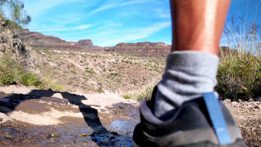 Low-angle view of a man with a backpack hiking on a sunny day along a rocky mountain path with a small stream, exploring the beautiful volcanic landscape of Gran Canaria, Canary Islands