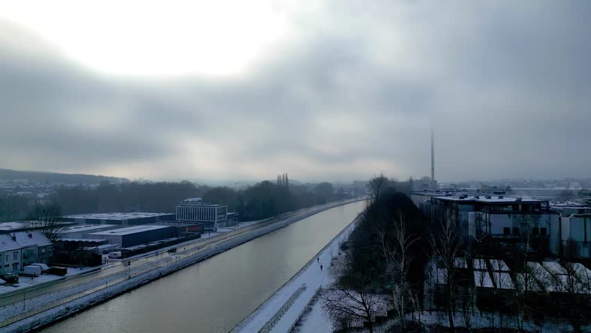 Halle, Vlaams Brabant, Belgium, 13th of January, 2026, Aerial winter view of a canal through an industrial cityscape, snowy banks, overcast sky and muted light, with buildings and trees along the waterway.