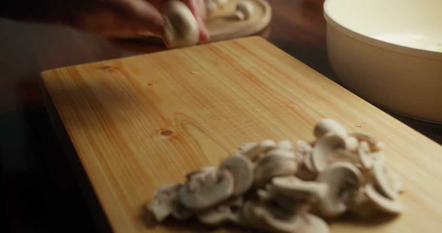 Close-up of a chef slicing fresh white mushrooms on a wooden cutting board. Hands preparing mushrooms with a kitchen knife for homemade cooking in warm natural light, rustic kitchen atmosphere.
