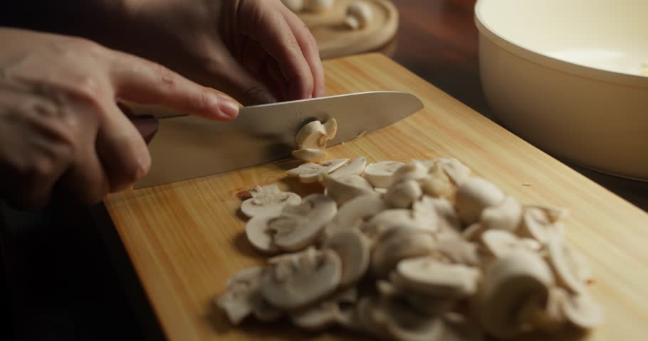 Close-up of a chef slicing fresh white mushrooms on a wooden cutting board. Hands preparing mushrooms with a kitchen knife for homemade cooking in warm natural light, rustic kitchen atmosphere.