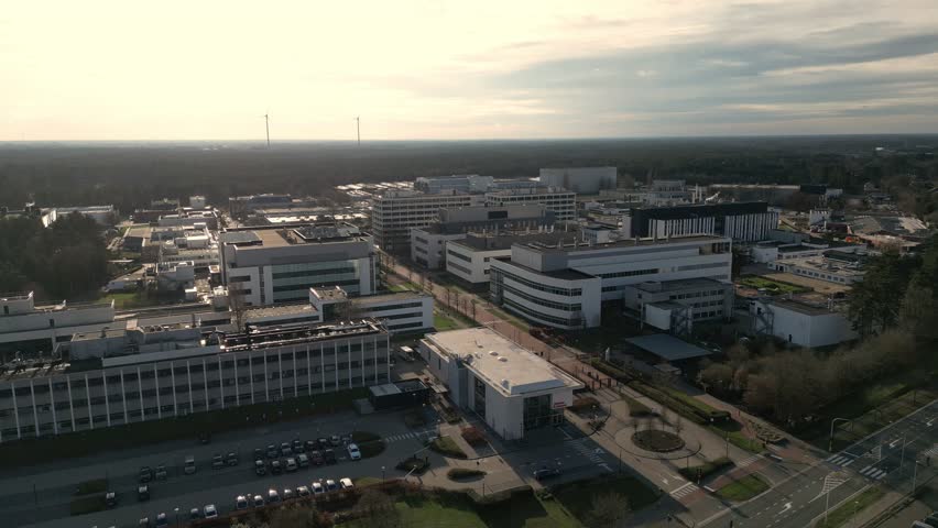 Aerial view of modern industrial or office campus with multiple buildings, roads and parking lots at sunset, surrounded by trees under cloudy sky.