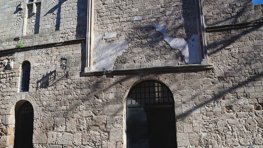 Slow upward pan revealing the facade and stone details of a medieval building in the town of Rhodes, Greece under clear blue sky