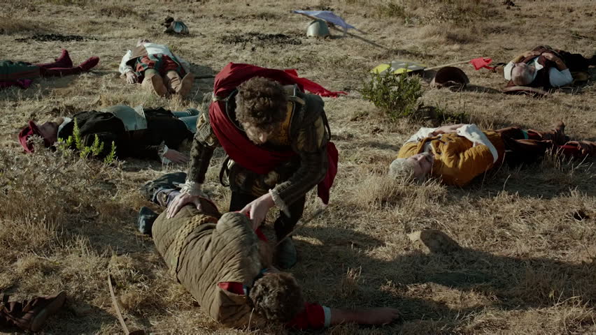 In a large open field, soldiers tend to fallen comrades after a battle. Worn armor is scattered, and the men share moments of grief and reflection during the day.