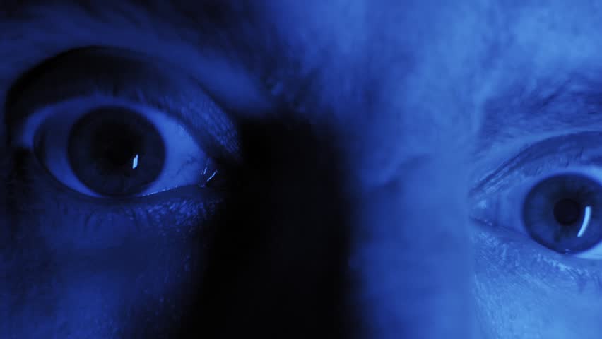Male face stares into camera with wide terrified eyes under monochromatic blue light. Extreme close up captures rapid blinking and intense paranoid expression in darkness.
