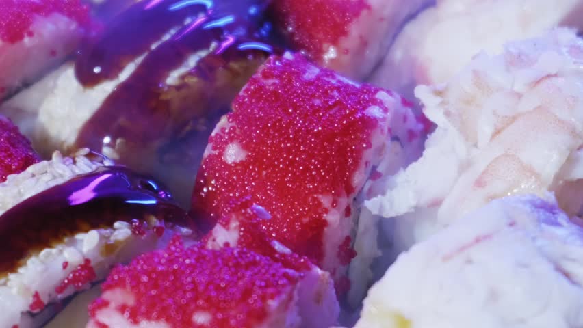 Wooden chopsticks grab a sushi roll covered in red fish roe from a crowded platter. Cool purple lighting highlights the texture of the rice and creamy filling.