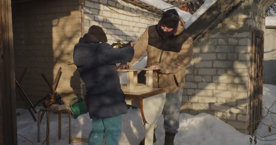 Young son learning from his father how to use a power drill while building a wooden bird feeder together in the backyard during a sunny winter day, fostering family bonding and practical skills.