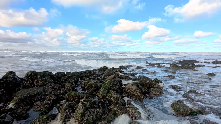 Rocky shoreline with moss-covered boulders, ocean waves and foam under bright blue sky with scattered clouds; wide coastal seascape background.