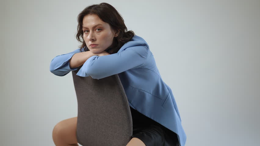 Stylish young woman in a blue jacket sits backwards on a chair. She leans forward, looking intently at the camera in a modern studio