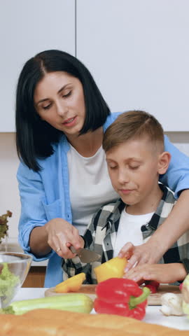 Good-looking satisfied young dark-haired mom teaching her 12-aged son to use the knife to cut yellow pepper during preparing tasty vegetable salad together with her husband in cozy cuisine