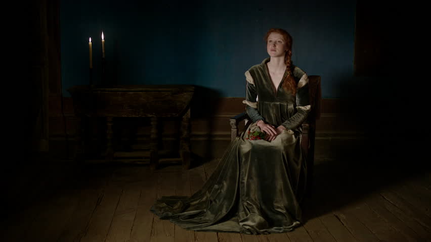 A woman wearing a green dress is seated alone in a dim room illuminated by candles. She appears deep in thought while gazing into the distance.