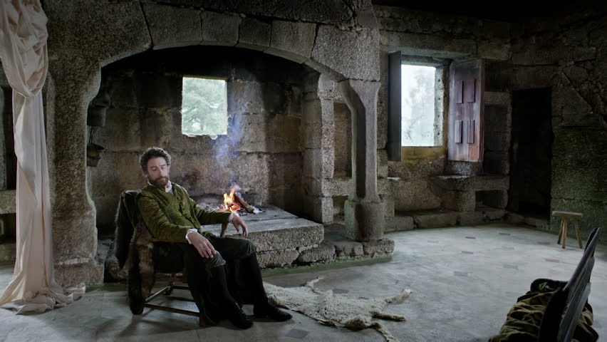 Two men meet in a cold stone room to discuss a serious topic. A fire burns in the middle of the room as they sit and stand by it, deep in thought.