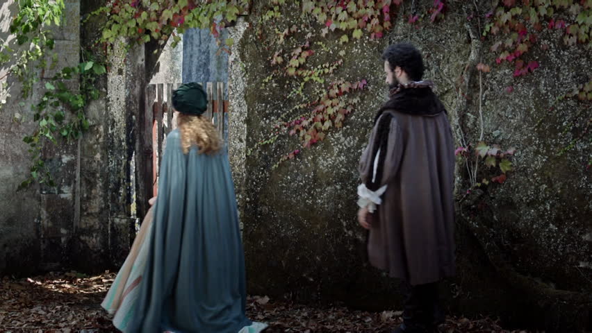 A couple walks through a stone wall covered in vines. They stop to look at a wooden gate. The leaves on the ground show that it is autumn. The setting feels old and mysterious.