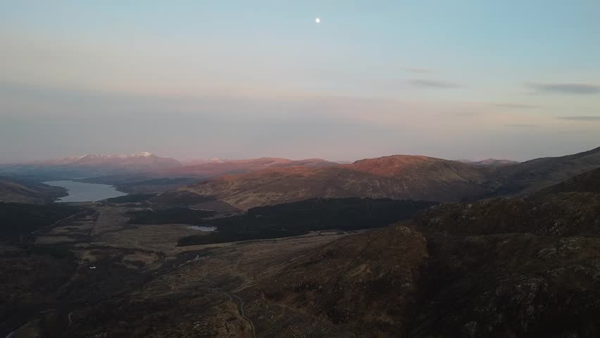 Aerial drone footage panning left to right over a long mountain loch at dusk, with rugged Highland hills catching soft evening light and calm water winding through the valley in peaceful conditions.