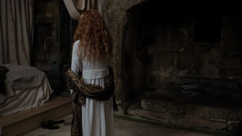 A woman wearing a white dress looks around an old room with a large fireplace. She seems curious about her surroundings while holding pieces of clothing. The atmosphere appears mysterious.