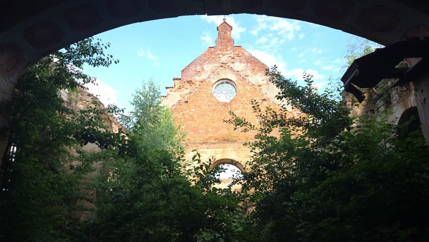 Abandoned red brick church ruins with an arched entrance and circular window sit overgrown with green foliage and trees under a clear blue sky, capturing a sense of lost history
