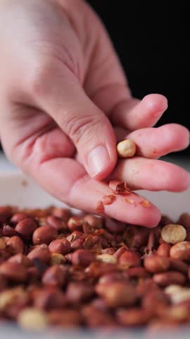 A close-up of a hand peeling roasted peanuts, with skins falling into a bowl of nuts