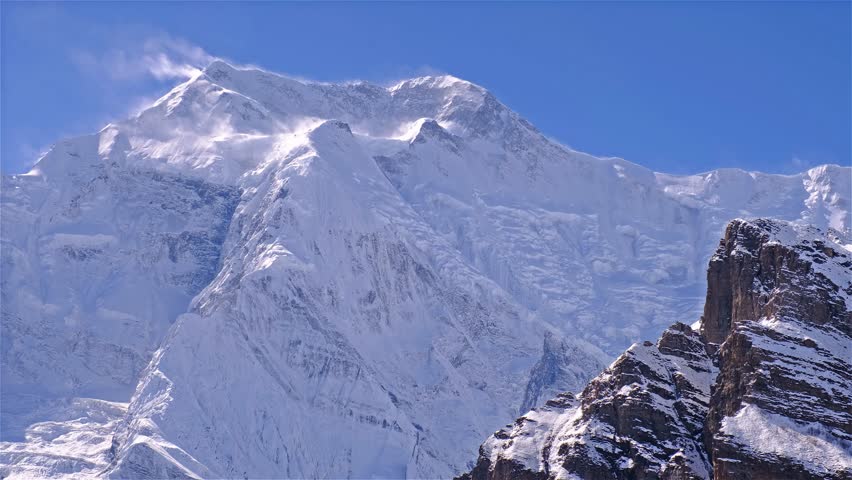Timelapse of the massive rock and ice wall of Annapurna II peak (7937 meters) after a recent snowfall. Hurricane-force winds strip snow from the steep face, forming dramatic snow plumes near the summit, often called snow flags. Extreme high-altitude conditions, powerful wind, and blowing snow emphasize the raw scale and intensity of the Himalayan mountain environment
